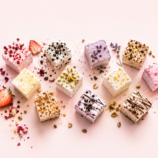 An overhead flat-lay of ten small square dessert pieces in assorted flavors, decorated with dried berries, chocolate chips, pistachios, zest, and drizzles, arranged on a soft pink background with scattered edible garnishes.